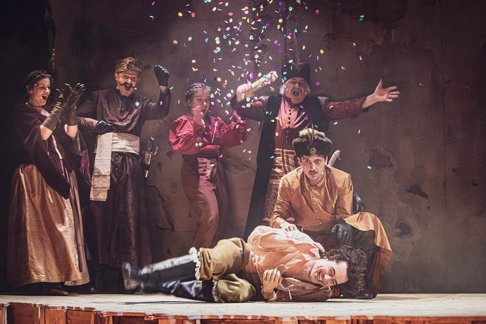 zdjęcie ze spektaklu "Pan Wołodyjowski". aktorzy ubrani z żupany, stroje historyczne, na pierwszym planie leży uśmiechnięty aktor, wszyscy uśmiechnięci, puszczają konfetti