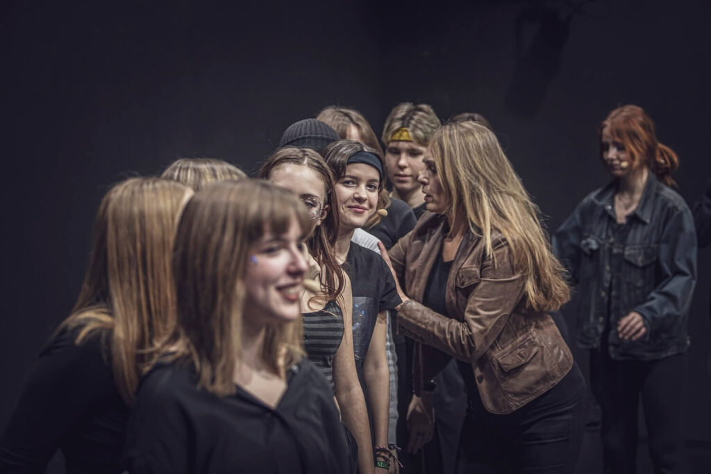 Zdjęcie z pierwszej edycji Festiwalu Otwarcia. Grupa młodzieży w kostiumach stoi w rzędzie na scenie. Obok nich stoi kobieta i trzyma jedną osobę za ramiona.