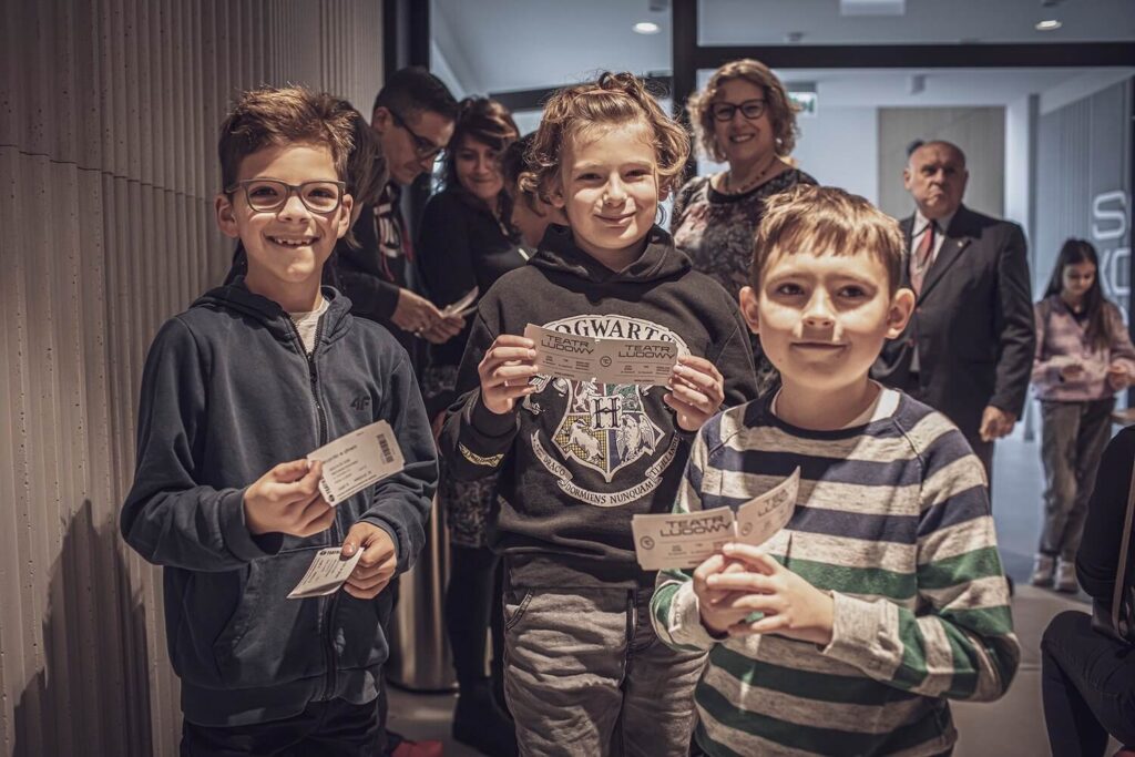 Zdjęcie z pierwszej edycji Festiwalu Otwarcia. Na pierwszym planie trzej chłopcy trzymają bilety do teatru i się uśmiechają. Za nimi stoi grupa dorosłych.