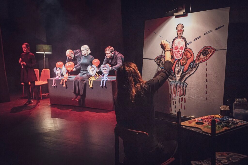 Zdjęcie z pierwszej edycji Festiwalu Otwarcia. Na scenie artystka maluje surrealistyczny obraz. W tle aktorzy i aktorka animują lalki teatralne z przyklejonymi twarzami mężczyzn.
