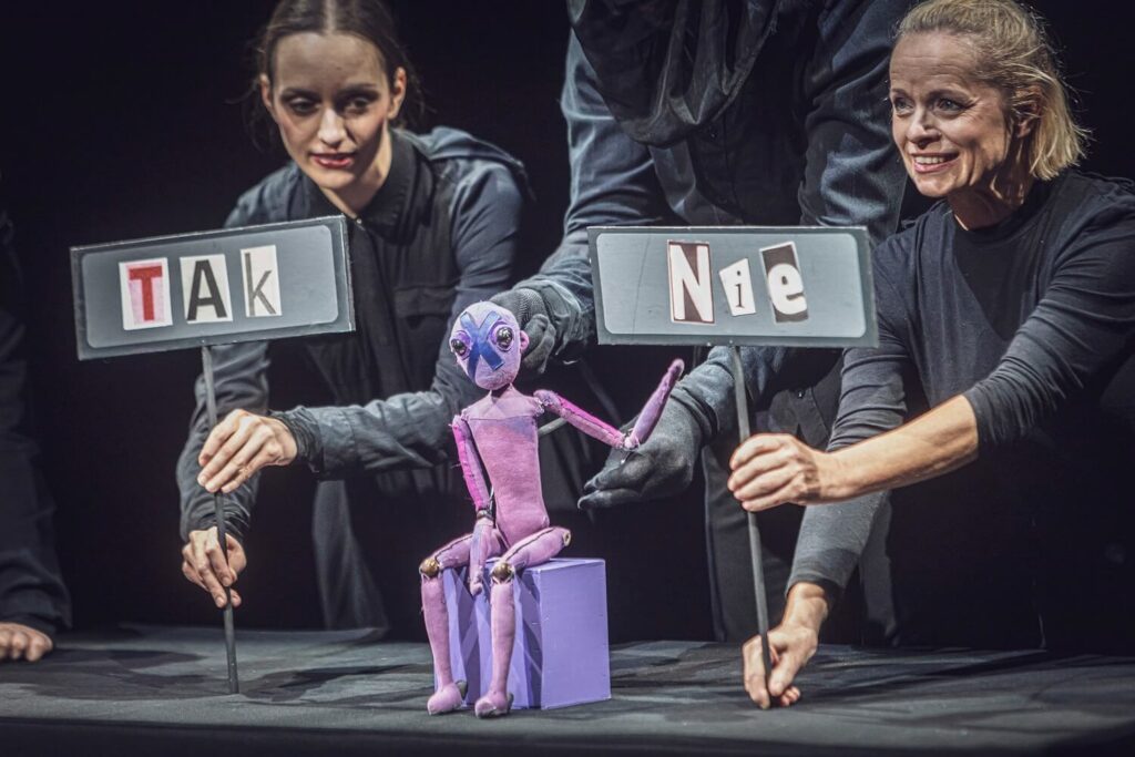 Zdjęcie z pierwszej edycji Festiwalu Otwarcia. Lalka teatralna siedzi na małym podeście. Jest trzymana przez osobę w rękawiczkach; obok aktorki pokazują tabliczki z napisami „Tak” i „Nie”.
