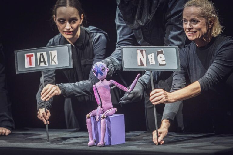 Zdjęcie z pierwszej edycji Festiwalu Otwarcia. Lalka teatralna siedzi na małym podeście. Jest trzymana przez osobę w rękawiczkach; obok aktorki pokazują tabliczki z napisami „Tak” i „Nie”.