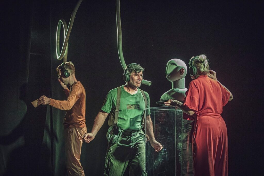Zdjęcie z pierwszej edycji Festiwalu Otwarcia. Dwóch aktorów i aktorka na scenie. Mają kolorowe stroje i słuchawki na uszach. Aktor i aktorka stoją przy szklanym postumencie i patrzą na siebie. Drugi aktor jest z tyłu. Nad nimi zwisa zwinięta rura.