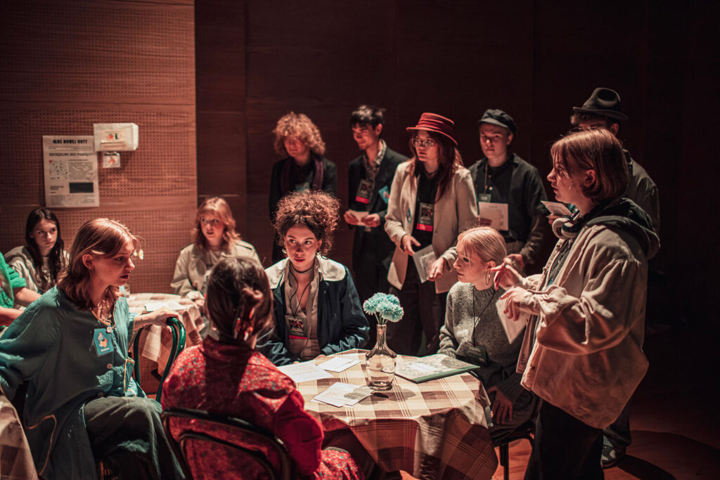 Zdjęcie z drugiej edycji Festiwalu Otwarcia. Gra teatralna. Młode dziewczyny siedzą z kartkami przy okrągłych stolikach. Wokół nich stoi grupa uczestników i uczestniczek z kartkami.