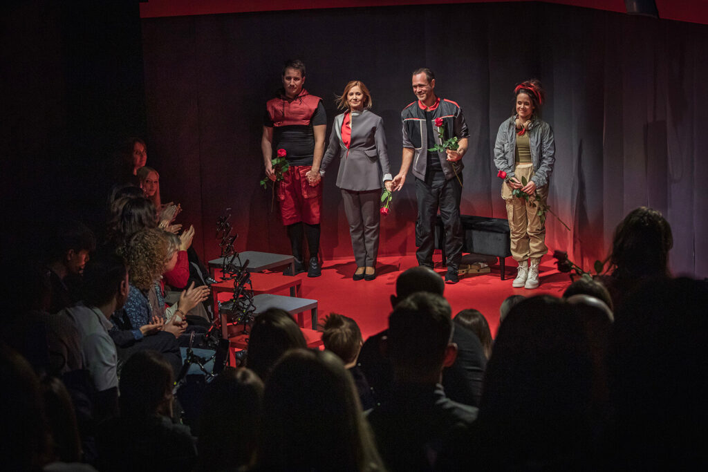 Zdjęcie z drugiej edycji Festiwalu Otwarcia. Na małej scenie stoją dwie aktorki i dwóch aktorów. Mają kostiumy z czerwonymi elementami, trzymają róże. Przed nimi publiczność siedzi i klaszcze.
