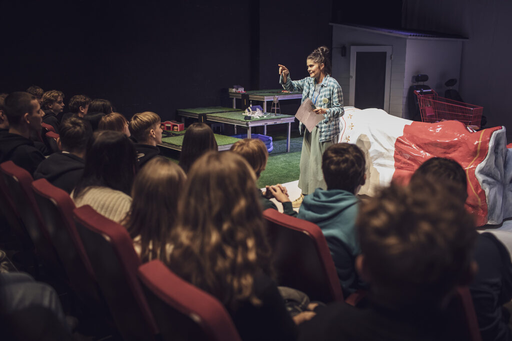 Zdjęcie z trzeciej edycji Festiwalu Otwarcia. Na scenie stoi kobieta, trzyma kartkę i wskazuje przed siebie. Przed nią na widowni siedzi grupa młodzieży. W tle jest scenografia: kanapa w kształcie puszki, wózek sklepowy, trawa, drzwi.