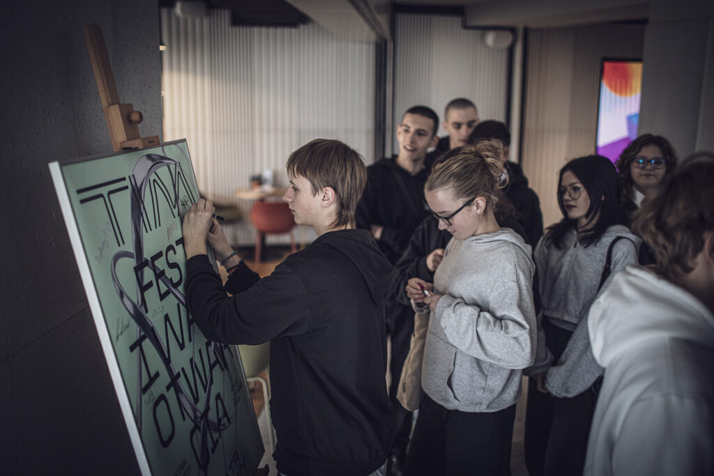 Zdjęcie z trzeciej edycji Festiwalu Otwarcia. Grupa młodzieży stoi przed plakatem Festiwalu Otwarcia. Trzymają markery. Jedna osoba podpisuje się na plakacie.