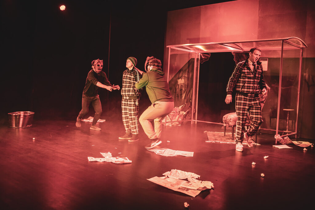 Zdjęcie z trzeciej edycji Festiwalu Otwarcia. Na scenie dwóch aktorów w maskach szarpie aktorkę w dresie w kratkę i czapce. Z boku idzie aktor w dresie w kratkę. Na scenie są rozrzucone gazety, z tyłu jest przystanek autobusowy. Zdjęcie zabarwione na kolor czerwony