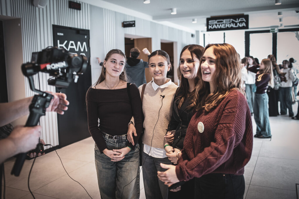 Zdjęcie z trzeciej edycji Festiwalu Otwarcia. Na korytarzu trzy dziewczyny stoją przed kamerą. W tle przy napisie „Scena kameralna” stoi młodzież.