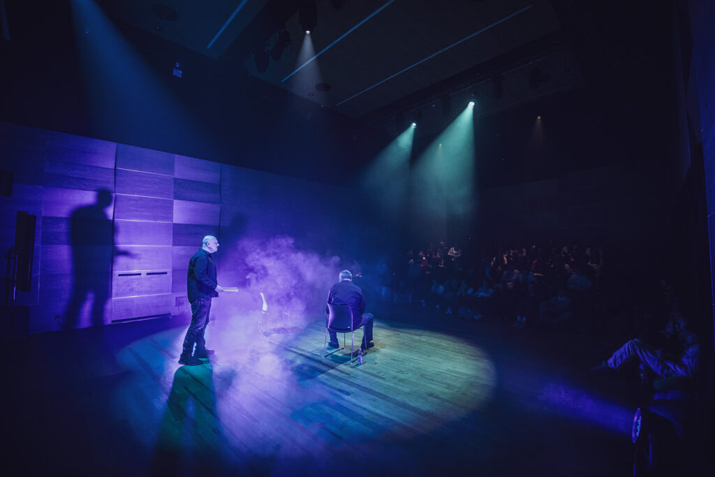Zdjęcie z trzeciej edycji Festiwalu Otwarcia. Na środku sceny jeden aktor siedzi tyłem, drugi stoi ze scenariuszem, wokół dym, nad nimi punktowe światło, przed nimi widownia. Zdjęcie zabarwione na kolor niebieski.