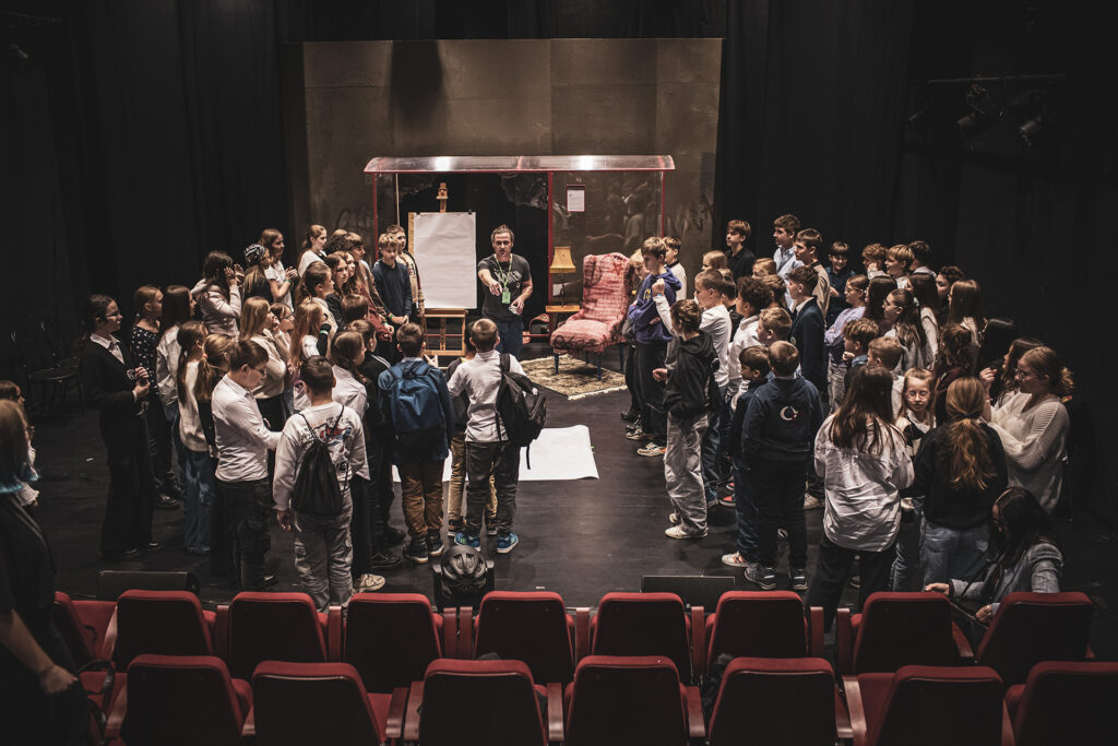 Zdjęcie z trzeciej edycji Festiwalu Otwarcia. Duża grupa dzieci na scenie. Mężczyzna z identyfikatorem Festiwalu stoi na środku przy sztaludze z dużą kartką. Z tyłu jest scenografia przedstawiająca przystanek autobusowy.