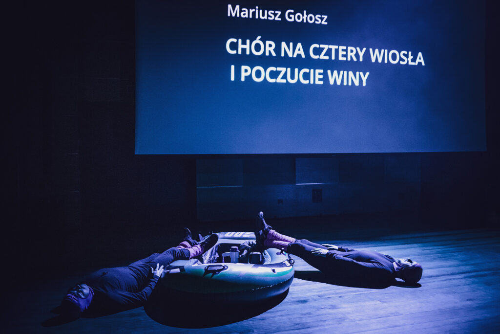 Zdjęcie z trzeciej edycji Festiwalu Otwarcia. Na scenie przy dmuchanym pontonie leży dwóch aktorów w kominiarkach i getrach. Nad nimi jest ekran z napisem „Mariusz Gołosz Chór na cztery wiosła i poczucie winy”.