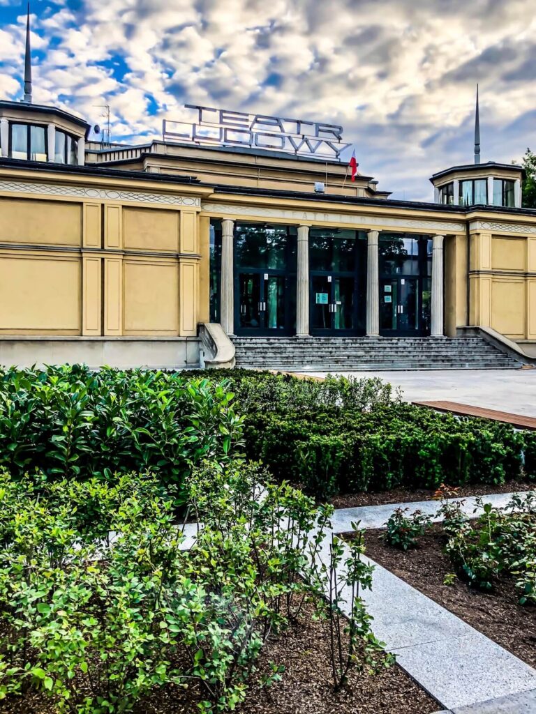 Widok na budynek główny teatru. Beżowy parterowy budynek o bryłowej konstrukcji. Po środku przeszklone wejście nad nim duży neon z napisem teatr ludowy, po bokach dwie przeszklone wierszyczki/ świetliki. Przed budynkiem chodnik i zieleniec.