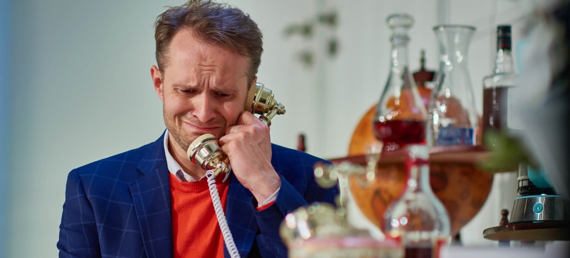 Zdjęcie ze spektaklu Kolacja dla głupca. Zbliżenie. Aktor w swetrze i marynarce w kratkę siedzi przy stole. Przy uchu trzyma słuchawkę starodawnego telefonu. Jest smutny.