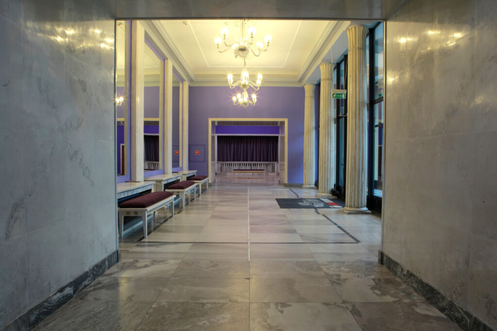 Widok foyer teatralnego, długi korytarz z marmurami na podłogach i ścianach. Po prawej stronie przeszklone wejście do budynku, po lewej wysokie do sufitu lustra.