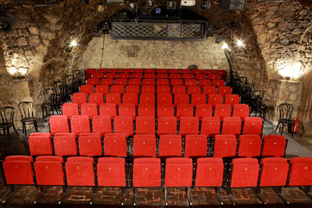Widok na widownie Sceny Pod Ratuszem, kamienne ściany z zaokrągleniem przy suficie. na pierwszym planie rzędy czerwonych foteli teatralnych.